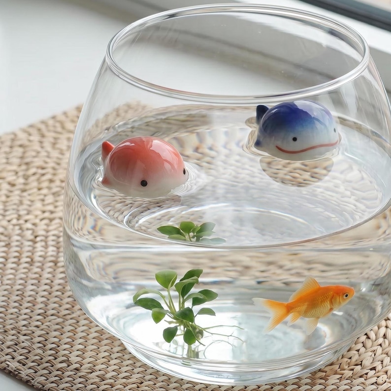 May include: A clear glass bowl filled with water containing a small orange goldfish, a green aquatic plant, and two small ceramic whale figurines, one colored in shades of pink and white and the other in shades of blue and white, floating on the surface.