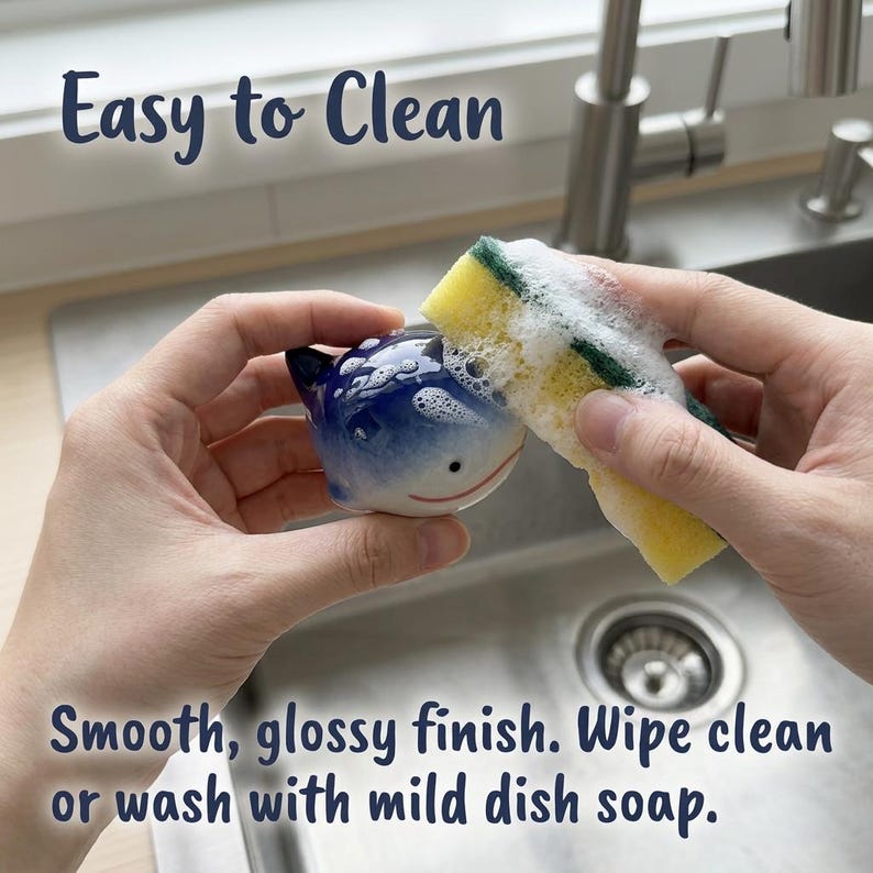 May include: A small, blue and white ceramic whale figurine with a smooth, glossy finish. The text "Easy to Clean" and "Smooth, glossy finish. Wipe clean or wash with mild dish soap." is visible. A yellow and green sponge is cleaning the whale.