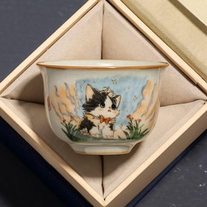 May include: A ceramic teacup with a hand-painted illustration of a calico kitten wearing a bow tie, set against a whimsical sky and floral background. The cup is presented in a wooden gift box with a soft, beige interior.