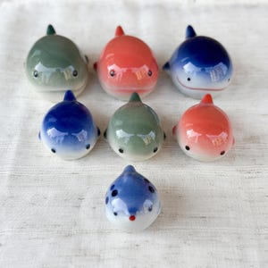 May include: Seven small ceramic whale figurines arranged in a group. The collection includes two green, two orange, and three blue whales, each featuring a smooth, glossy finish, rounded bodies, small fins, and simple painted eyes and mouths.