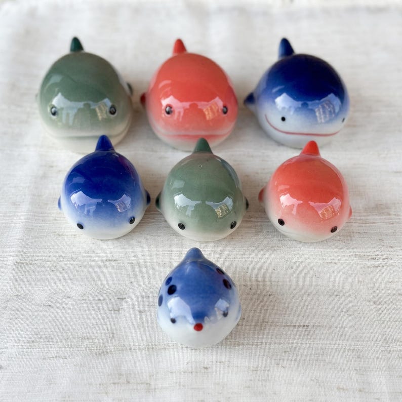 May include: Seven small ceramic whale figurines arranged in a group. The collection includes two green, two orange, and three blue whales, each featuring a smooth, glossy finish, rounded bodies, small fins, and simple painted eyes and mouths.