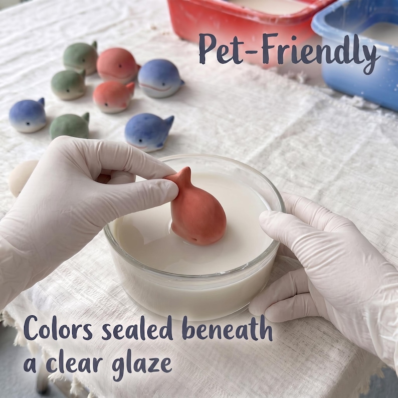 May include: Small ceramic whale figurines in shades of blue, green, and coral are arranged on a white cloth. One coral whale is being dipped into a clear glass bowl filled with a white liquid glaze. Text reads Pet-Friendly and Colors sealed beneath a clear glaze.
