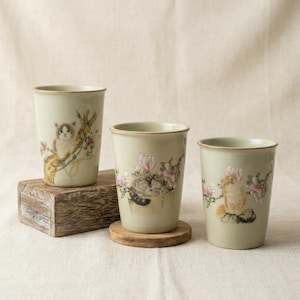 May include: Three light green ceramic cups featuring hand-painted illustrations of cats nestled among pink flowers and branches. Each cup has a smooth, cylindrical shape with a slightly flared rim, showcasing detailed feline art in soft tones.