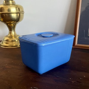 May include: A rectangular, light blue ceramic butter dish with a matching lid. The lid has a small, round handle and a series of curved lines. The dish is on a dark wooden surface, with a brass lamp and framed picture in the background.