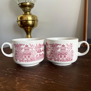 May include: A pair of white ceramic mugs featuring a red and white Asian-inspired pattern. Each mug has a handle and a detailed illustration of pagodas and trees. The mugs are on a wooden surface.
