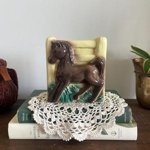 May include: A ceramic horse figurine featuring a brown horse standing in front of a pale yellow fence and green grass. The rectangular piece has a glossy finish and depicts the horse in profile, facing left, with a raised mane and tail.