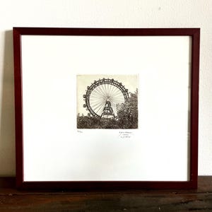 May include: A framed black and white etching of the Wiener Riesenrad Ferris wheel in Vienna. The artwork is centred on a white mat within a dark brown wooden frame. Below the image, the text reads 14/100, 1992, and WIEN in pencil.