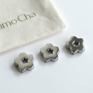 May include: Three silver flower-shaped objects with a star-shaped center are displayed next to a small, off-white bag. The bag has the text "KumoCha" printed on it. A green ribbon is visible in the background.