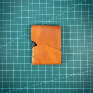 May include: A tan leather card holder featuring a diagonal front pocket design and white contrast stitching along the left side. The wallet has a smooth, matte texture and a rectangular shape with a cutout section on the upper left edge.