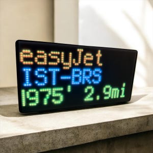 Puede incluir: Una pantalla digital negra con el texto "easyJet IST-BRS 1975' 2.9mi" en luces LED naranjas, azules y verdes. La pantalla es rectangular con esquinas redondeadas y está sobre una superficie gris.