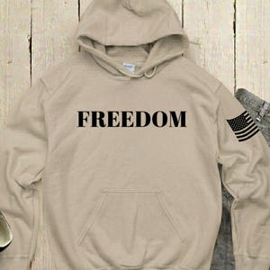 May include: A beige hooded sweatshirt with the word "FREEDOM" printed in black across the chest. An American flag is on the left sleeve. The sweatshirt has a front pocket and a drawstring hood.