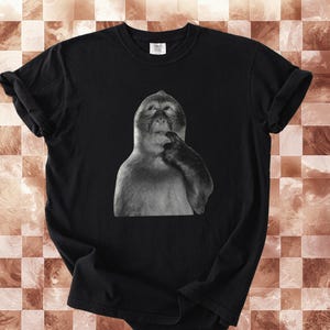 May include: Black t-shirt featuring a grayscale image of a monkey with its finger to its mouth, as if deep in thought. The shirt is made of a soft material and has a classic crew neck design. The image is centered on the front of the shirt.