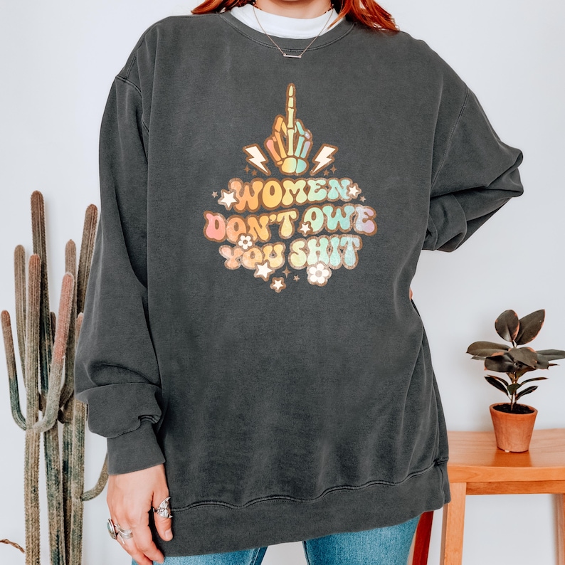 May include: Dark gray sweatshirt with the text "WOMEN DON'T OWE YOU SHIT" in a retro font. The design includes a hand giving the middle finger, lightning bolts, and floral accents. The sweatshirt is a relaxed fit.