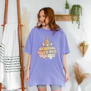May include: A periwinkle-colored t-shirt with the text "WOMEN DON'T OWE YOU SHIT" in a retro font. The graphic includes a hand giving the middle finger, lightning bolts, and flowers.