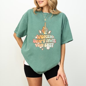 May include: Sage green t-shirt with the text "WOMEN DON'T OWE YOU SHIT" in a retro font. The graphic includes a hand giving the middle finger, lightning bolts, stars, and flowers. The shirt is a short-sleeved, crew neck style.