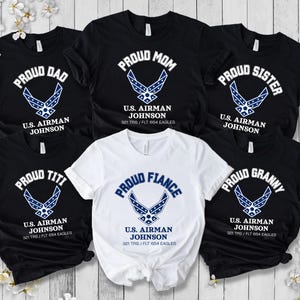 May include: A collection of black and white t-shirts with the text "Proud Dad", "Proud Mom", "Proud Sister", "Proud Titi", "Proud Fiance", and "Proud Granny". Each shirt features a blue and white emblem and the text "U.S. Airman Johnson".