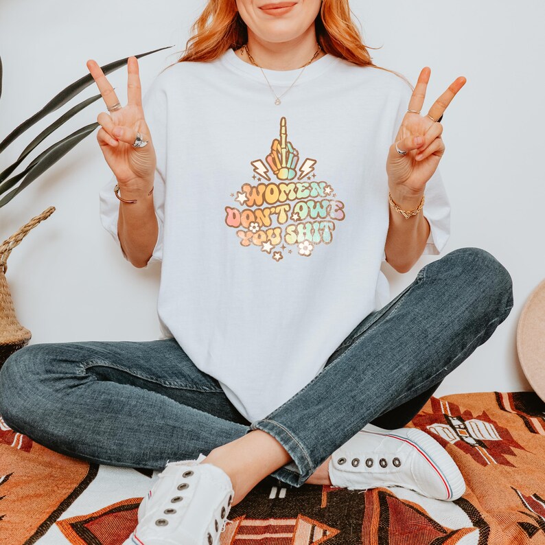 May include: White t-shirt with the text "WOMEN DON'T OWE YOU SHIT" in a retro font, featuring a middle finger graphic. The shirt is paired with blue jeans and white sneakers. The person is sitting cross-legged.