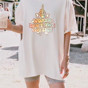 May include: Cream-colored t-shirt with the text "WOMEN DON'T OWE YOU SHIT" in a colorful, retro font. The design includes a hand giving the middle finger. The shirt is a relaxed fit.