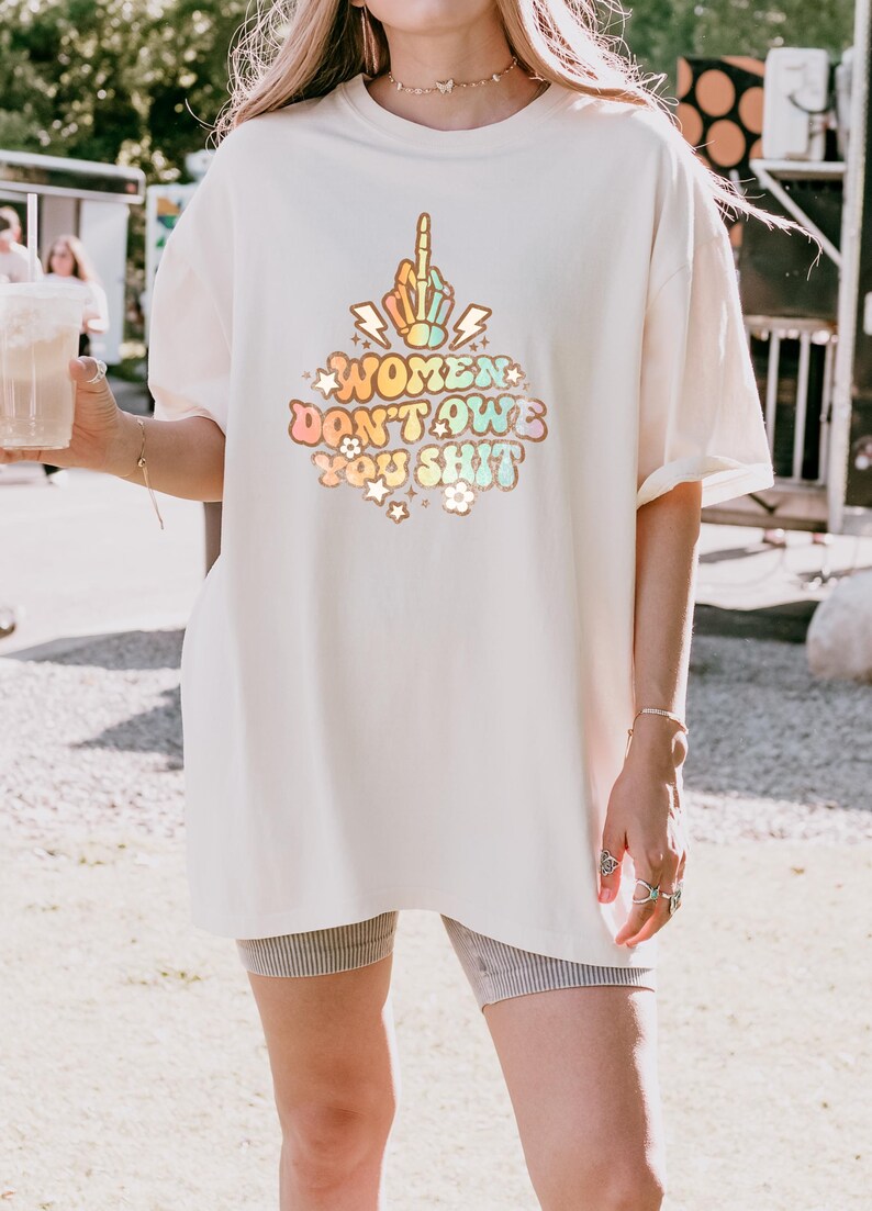 May include: Cream-colored t-shirt with the text "WOMEN DON'T OWE YOU SHIT" in a colorful, retro font. The design includes a hand giving the middle finger. The shirt is a relaxed fit.