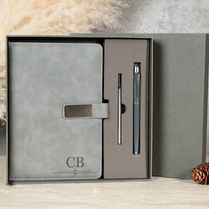 Puede incluir: Un juego de regalo que incluye un diario de cuero gris con un cierre metálico y las iniciales "CB" grabadas en la portada. El conjunto también incluye un bolígrafo y una recarga, todo en una caja gris.