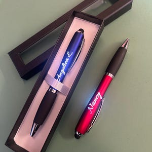 May include: Two personalized pens, one blue and one red, displayed in black gift boxes. The blue pen is inscribed with "Angelica L", and the red pen reads "Nancy". Both pens have black grips and silver accents, ideal for gifts or personal use.