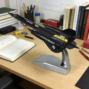 Puede incluir: Maqueta negra de avión SR-71 Blackbird con inscripciones "U.S. AIR FORCE" y "NASA". El modelo está montado sobre un soporte plateado y se exhibe sobre un escritorio de madera. El modelo tiene detalles en amarillo y rojo.