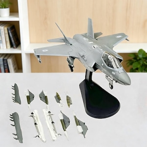 May include: A gray model airplane with a black stand. The model has detailed wings, a cockpit, and landing gear. Several detachable components are arranged in front of the plane. The model is a detailed replica of a fighter jet.