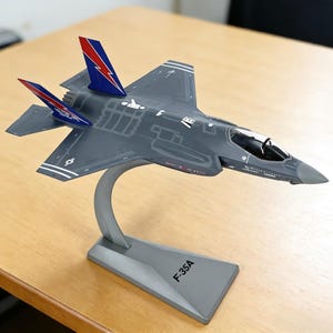 May include: A gray F-35A model airplane with red and blue accents on the tail fins, mounted on a silver stand. The model has detailed markings and is displayed on a wooden surface.