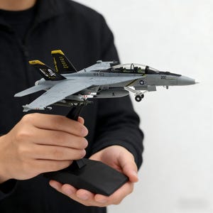 Op de afbeelding: Een grijs en zwart modelvliegtuig met de tekst "VFA-103" en "200" op de romp. Het model heeft gele accenten op de staart en is gemonteerd op een zwarte standaard. Het model wordt door een persoon vastgehouden.