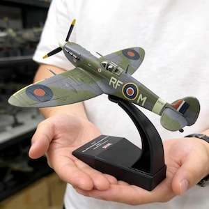 May include: A detailed model of a WWII-era fighter plane, featuring olive green and gray camouflage. The model has a propeller with yellow tips, roundels, and the markings "RF O M". It is mounted on a black display stand.