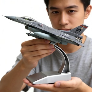 以下が含まれることがあります： ライトブルーの着陸装置とシルバーのディスプレイスタンドを備えた、グレーのF-16ファイティングファルコンモデル飛行機。モデルには詳細なマーキングがあり、人の前に保持されています。モデルの長さは約25cmです。