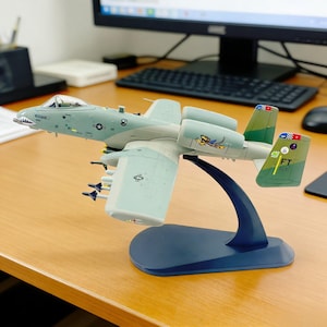 Puede incluir: Maqueta de un avión A-10 Thunderbolt II, pintado en tonos verdes y grises, con un diseño de boca de tiburón en la nariz. El modelo está montado sobre un soporte azul, mostrando los detalles de las alas y la cola.