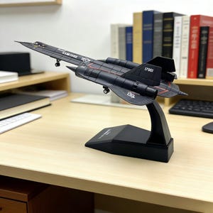 Puede incluir: Maqueta negra de avión SR-71 Blackbird con líneas rojas y la inscripción "U.S. AIR FORCE". El modelo está montado sobre un soporte negro y expuesto sobre un escritorio de madera.