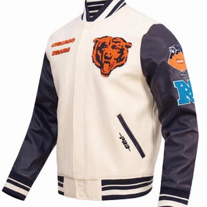 May include: A cream-colored varsity jacket with navy blue sleeves, featuring the Chicago Bears text and a bear head logo on the chest. The sleeve displays a football graphic and a large letter N. It has a snap-button closure and striped ribbed trim.