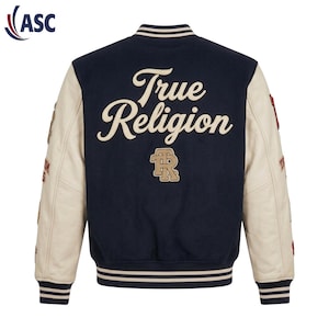 Könnte beinhalten: Eine marineblaue Collegejacke mit cremefarbenen Ärmeln. Auf der Rückseite befindet sich der Schriftzug True Religion in cremefarbener Schreibschrift, ein beiges TR-Logo sowie gestreifte Rippbündchen an Kragen, Ärmeln und Saum. Patches auf den Ärmeln.