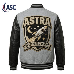 Könnte beinhalten: Eine graue Collegejacke mit schwarzen Lederärmeln und gestreiften Rippbündchen. Auf der Rückseite befindet sich ein großer gestickter Aufnäher mit einer Rakete, einem Mond und Sternen sowie dem Schriftzug ASTRA EXPLORERS' GUILD in Schildform.