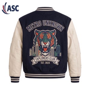 Könnte beinhalten: Eine marineblaue Collegejacke mit cremefarbenen Ärmeln und gestreiften Rippbündchen. Auf der Rückseite befindet sich eine gestickte Tigerkopf-Grafik über einer Stadtsilhouette mit dem Schriftzug METRO UNKNOWN, EXPLORERS CLUB und EST. 2024 in Weiß.