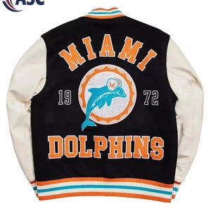 May include: A black varsity jacket with cream-coloured sleeves, featuring the text MIAMI DOLPHINS in orange, the numbers 19 and 72, and a central graphic of a teal dolphin wearing a helmet inside an orange sunburst circle, with striped trim.