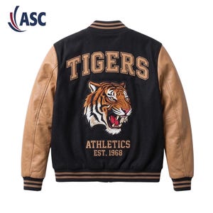 May include: A black varsity jacket with tan leather sleeves, featuring a large embroidered tiger head on the back. The text TIGERS is arched above the tiger, with ATHLETICS EST. 1968 printed below. The collar, cuffs, and hem have black and tan stripes.