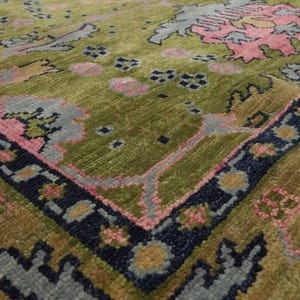 May include: Close-up of a section of a patterned rug. The rug features a green, pink, and blue floral design with a dark blue border. The rug's texture is visible, suggesting a woven construction.