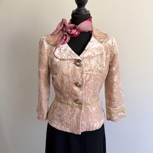 Puede incluir: Chaqueta vintage color rubor con estampado floral, ribete dorado y tres botones ornamentados. Una bufanda a juego con rayas rojas y blancas está drapeada alrededor del cuello del maniquí. La chaqueta se combina con una falda negra.