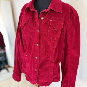 May include: A red corduroy jacket with a collared neckline, two chest pockets, and button closures. The jacket has a classic design with a slightly fitted silhouette. The fabric appears soft and has a subtle sheen. The jacket is displayed on a black mannequin.
