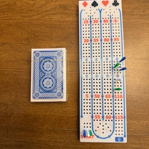 May include: A white plastic cribbage board with blue track lines, red numbers, and suit symbols at the top, featuring blue, green, and white pegs inserted into the holes. Beside it is a deck of playing cards with a blue and white geometric back.