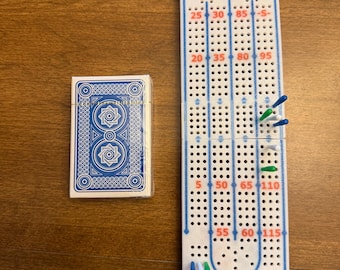 May include: A white plastic cribbage board with blue track lines, red numbers, and suit symbols at the top, featuring blue, green, and white pegs inserted into the holes. Beside it is a deck of playing cards with a blue and white geometric back.