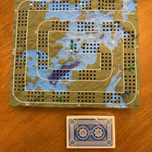 May include: A rectangular cribbage board featuring a topographical map design with blue water and green land, labeled with Thomas Lake, Fraser Lake, and Cacabic Lake. The board has numbered tracks and colored pegs, accompanied by a deck of blue playing cards.