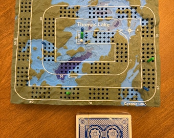 May include: A rectangular cribbage board featuring a topographical map design with blue water and green land, labeled with Thomas Lake, Fraser Lake, and Cacabic Lake. The board has numbered tracks and colored pegs, accompanied by a deck of blue playing cards.