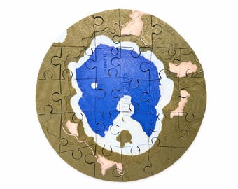 May include: A circular topographic puzzle of Crater Lake featuring a deep blue center representing the water, surrounded by white and tan land contours. The puzzle pieces are interlocking and include the text Crater Lake and 1947 R.