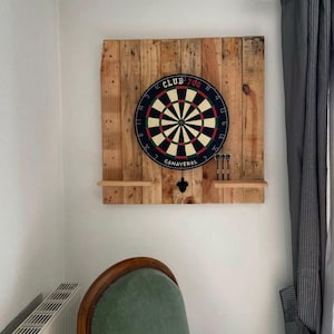 Puede incluir: Un tablero de dardos montado sobre un fondo de palet de madera. El tablero de dardos es negro, rojo y blanco con el texto "CLUB 700" y "CANAVERAL". Debajo del tablero de dardos hay dos pequeñas estanterías, con dardos y un soporte para dardos.