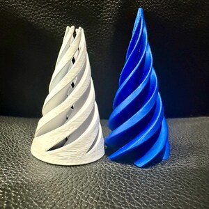 May include: Two spiral-shaped decorative objects. One is white, the other is blue. Both have a conical shape with a spiraling design. The objects are placed on a dark, textured surface. The design is reminiscent of a stylized horn or cone.