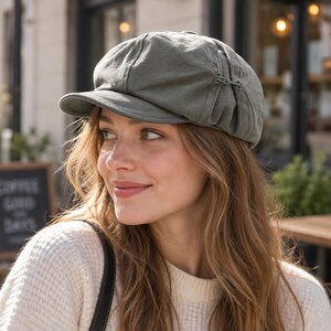 May include: An olive green newsboy cap featuring a structured brim and a small metallic decorative buckle detail on the side. The hat is crafted from a soft fabric with visible paneling and a rounded crown, providing a classic silhouette.